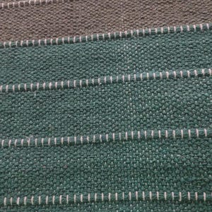 Op de afbeelding: Een geweven stof met een getextureerd oppervlak met horizontale strepen in diep bosgroen en gedempt olijfbruin. Het weefsel vertoont een consistent rasterpatroon met dunne, lichtgekleurde draadaccenten die horizontaal over het materiaal lopen.