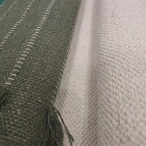 Op de afbeelding: Een getextureerd textiel met een donker olijfgroen gedeelte met verticale witte stiksels en gerafelde randen, naast een opgerold gedeelte van gebroken witte, grof geweven stof met een zichtbaar rasterachtig patroon over het geheel.