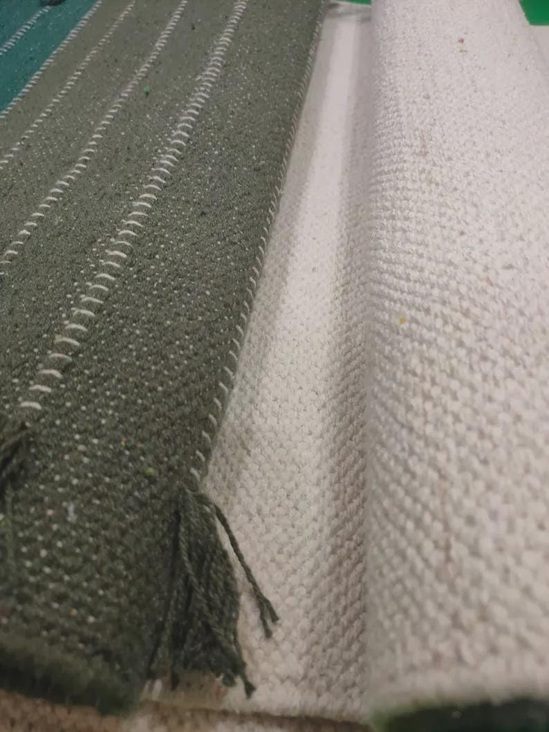 Op de afbeelding: Een getextureerd textiel met een donker olijfgroen gedeelte met verticale witte stiksels en gerafelde randen, naast een opgerold gedeelte van gebroken witte, grof geweven stof met een zichtbaar rasterachtig patroon over het geheel.