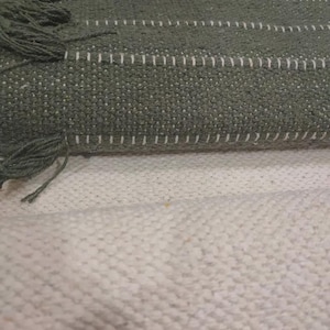 Op de afbeelding: Een geweven textiel met horizontale strepen in tinten donkergroen en olijf, geaccentueerd met dunne witte lijnen en afgewerkt met franjes. De stof vertoont een getextureerd, grof weefpatroon, gelaagd over een effen gebroken witte basis.