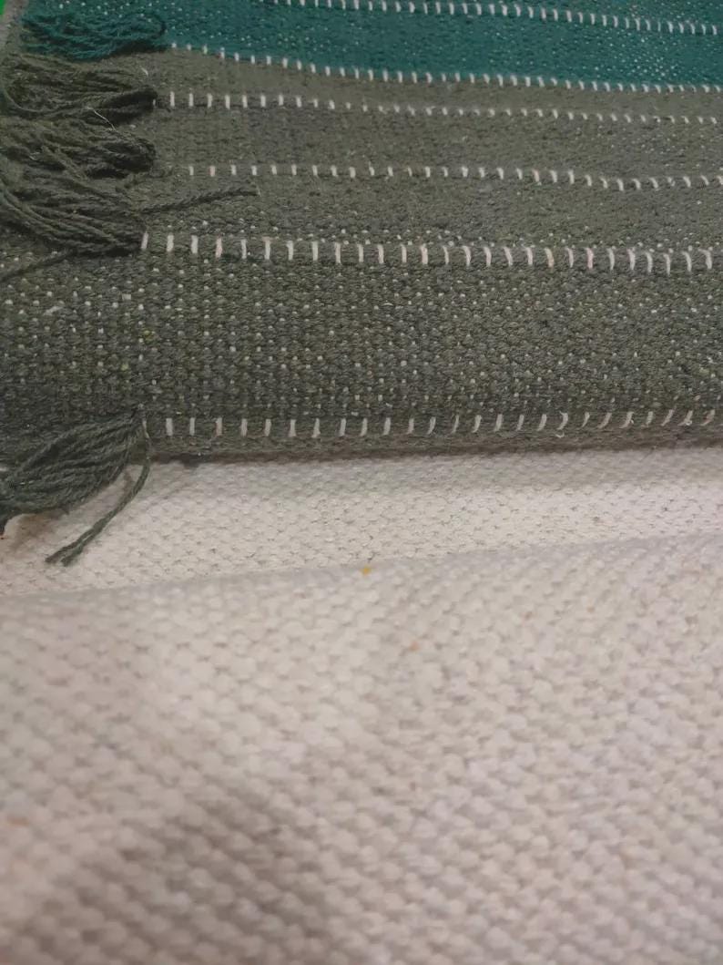 Op de afbeelding: Een geweven textiel met horizontale strepen in tinten donkergroen en olijf, geaccentueerd met dunne witte lijnen en afgewerkt met franjes. De stof vertoont een getextureerd, grof weefpatroon, gelaagd over een effen gebroken witte basis.