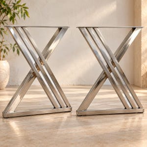 Puede incluir: Un par de patas de mesa de metal color plateado con un diseño en forma de X. Cada conjunto de patas consta de tres barras paralelas que se cruzan en el centro, conectadas por una barra base horizontal en la parte inferior y una placa de montaje plana arriba.