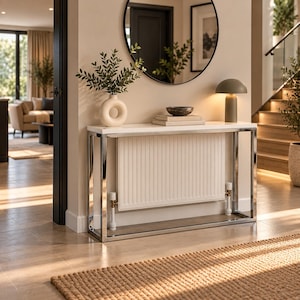 Puede incluir: Una mesa consola rectangular con superficie blanca y marco de metal cromado pulido. La mesa integra un radiador blanco de nervaduras verticales en el centro, flanqueado por dos válvulas blancas. Un jarrón blanco en forma de dona con plantas encima.