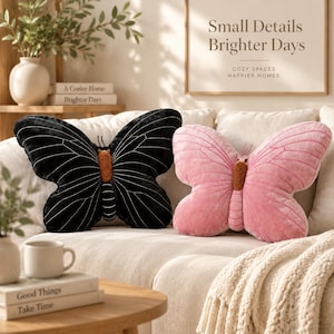 May include: Two butterfly-shaped throw pillows, one black with white embroidered wing veins and one pink with matching pink wing veins, both featuring a textured brown central body and small antennae, resting on a light-colored sofa.