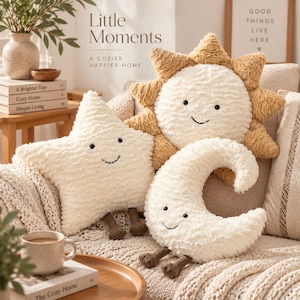 May include: Three plush pillows shaped like a sun, a star, and a crescent moon. Each pillow features a textured cream or tan fabric, a smiling face with black embroidered eyes and a mouth, and small brown dangling legs attached at the bottom.