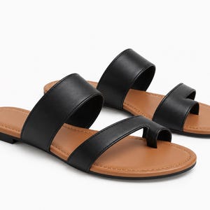 May include: A pair of flat sandals featuring two black straps across the top, with one strap designed as a toe loop. The footbed is a tan brown color with visible stitching along the edge, and the sole is black. The sandals are shown from a side angle.