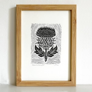 May include: A framed black and white linocut print of a thistle. The artwork shows a detailed depiction of the plant's flower and leaves, set against a textured background. The print is displayed in a natural wood frame.