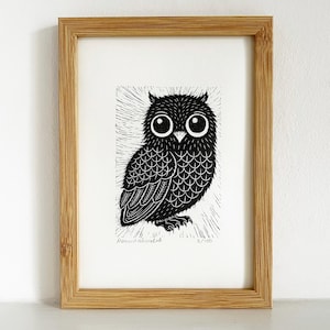 May include: Black and white owl print in a wooden frame. The owl has large, round eyes and detailed feather patterns. The print is numbered 2/100 and signed. The frame is a light brown colour.