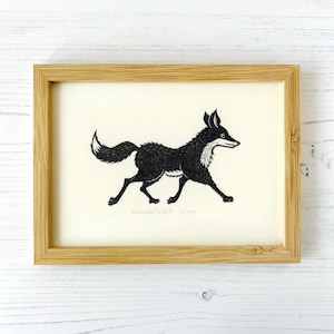 May include: A framed black and white linocut print featuring a stylised fox in a walking pose. The artwork is set against a cream-coloured background and enclosed in a light-toned wooden frame. The print includes handwritten text at the bottom.