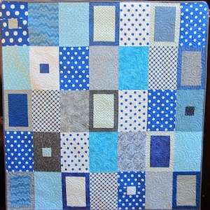 May include: A blue and white quilt with a patchwork design. The quilt is made up of squares of different fabrics, including polka dots, swirls, and solid colors. The quilt is edged in a gray fabric.