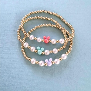 K&ouml;nnte beinhalten: Drei Armb&auml;nder mit goldfarbenen Perlen und wei&szlig;en Perlen. Jedes Armband hat einen andersfarbigen Mickey Mouse-f&ouml;rmigen Anh&auml;nger: rosa, t&uuml;rkis und lavendelfarben. Die Armb&auml;nder werden auf einer hellblauen Oberfl&auml;che pr&auml;sentiert.