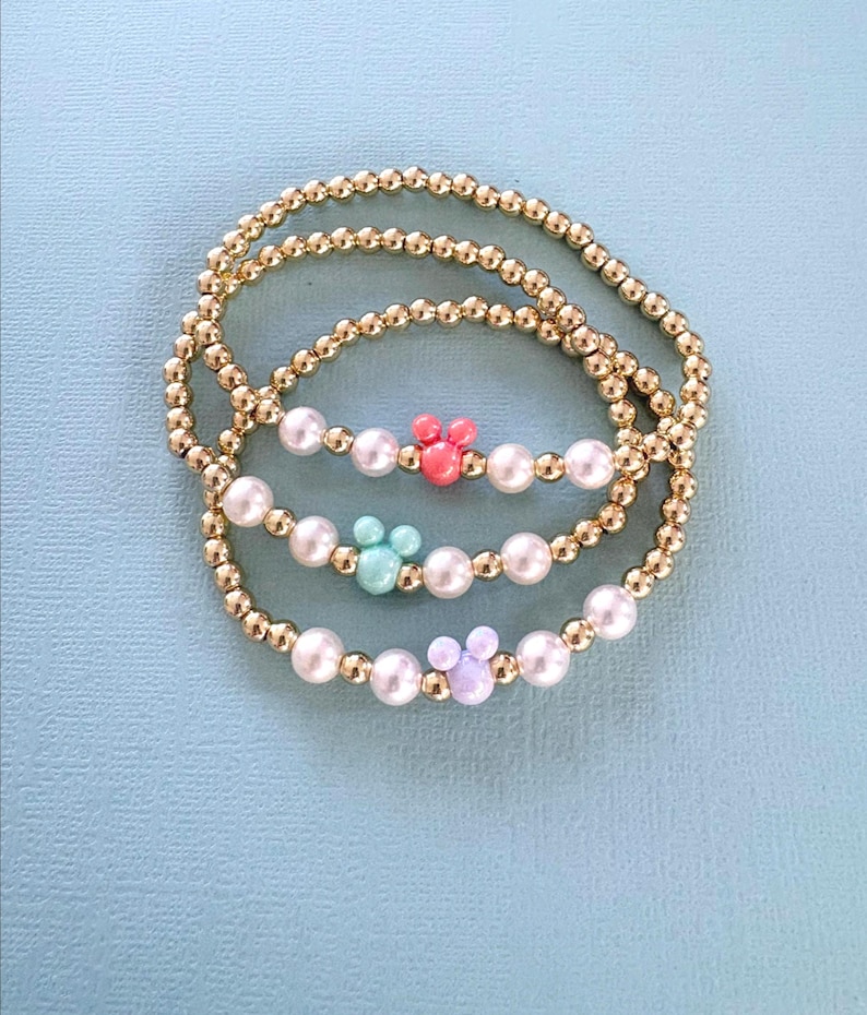 K&ouml;nnte beinhalten: Drei Armb&auml;nder mit goldfarbenen Perlen und wei&szlig;en Perlen. Jedes Armband hat einen andersfarbigen Mickey Mouse-f&ouml;rmigen Anh&auml;nger: rosa, t&uuml;rkis und lavendelfarben. Die Armb&auml;nder werden auf einer hellblauen Oberfl&auml;che pr&auml;sentiert.