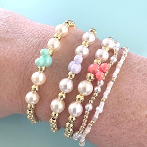 K&ouml;nnte beinhalten: F&uuml;nf Armb&auml;nder mit Perlen in verschiedenen Designs. Die Armb&auml;nder haben Kunstperlen, goldfarbene Perlen und Akzentperlen in Mintgr&uuml;n, Lavendel und Koralle. Die Armb&auml;nder werden an einem Handgelenk vor einem hellblauen Hintergrund getragen.