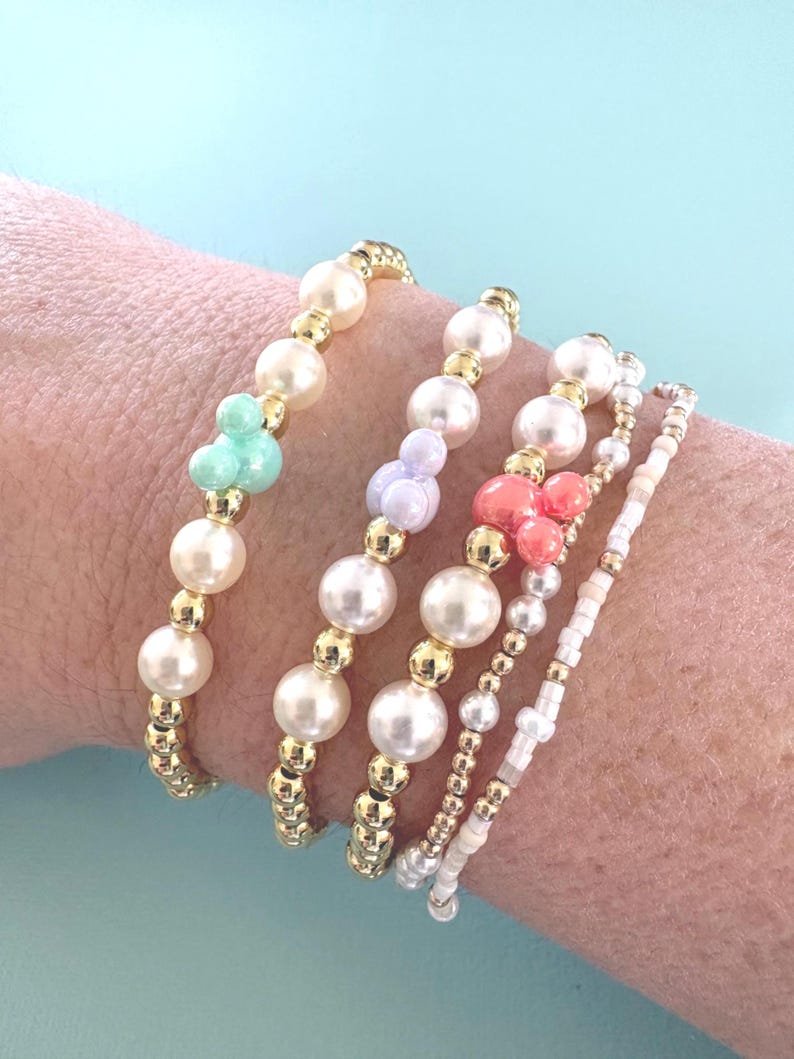 K&ouml;nnte beinhalten: F&uuml;nf Armb&auml;nder mit Perlen in verschiedenen Designs. Die Armb&auml;nder haben Kunstperlen, goldfarbene Perlen und Akzentperlen in Mintgr&uuml;n, Lavendel und Koralle. Die Armb&auml;nder werden an einem Handgelenk vor einem hellblauen Hintergrund getragen.