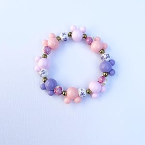 May include: A pink and purple beaded bracelet with gold accents and a Mickey Mouse head design. The beads are a mix of solid colors and floral patterns.
