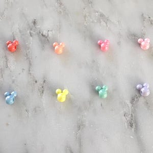 K&ouml;nnte beinhalten: Acht farbenfrohe, perlenartige Anh&auml;nger in Form eines M&auml;usekopfes sind auf einer Marmoroberfl&auml;che angeordnet. Die Anh&auml;nger sind in den Farbt&ouml;nen Rot, Orange, Rosa, Hellrosa, Blau, Gelb, Gr&uuml;n und Lavendel gehalten. Diese Anh&auml;nger k&ouml;nnten f&uuml;r die Schmuckherstellung oder andere Bastelarbeiten verwendet werden.