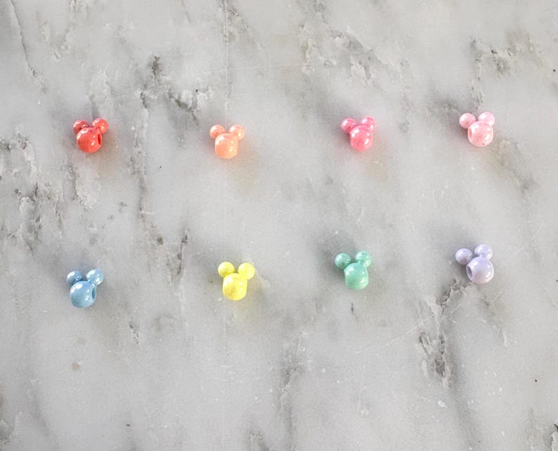 K&ouml;nnte beinhalten: Acht farbenfrohe, perlenartige Anh&auml;nger in Form eines M&auml;usekopfes sind auf einer Marmoroberfl&auml;che angeordnet. Die Anh&auml;nger sind in den Farbt&ouml;nen Rot, Orange, Rosa, Hellrosa, Blau, Gelb, Gr&uuml;n und Lavendel gehalten. Diese Anh&auml;nger k&ouml;nnten f&uuml;r die Schmuckherstellung oder andere Bastelarbeiten verwendet werden.