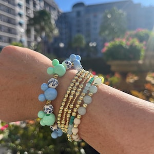 May include: A stack of bracelets with gold beads, light blue and green beads, and white beads with a blue and white floral pattern. The beads are arranged in a Mickey Mouse shape.