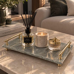 May include: A rectangular green stone tray with gold-toned metal handles, featuring a glass reed diffuser with black sticks, a cylindrical speckled candle, and a small glass jar with a gold lid, all arranged on the tray surface.