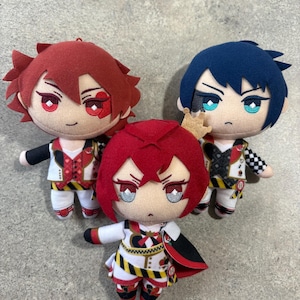 May include: Three plush dolls featuring anime-style characters. One doll has red hair and a heart mark, one has dark blue hair and teal eyes, and the centre doll wears a small gold crown, red hair, and a white, red, and black outfit.