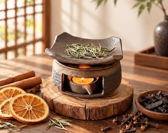 May include: A dark brown ceramic oil burner with a textured finish and the words good luck inscribed on the front. The burner features a top dish holding dried herbs and a lower chamber containing a lit tea light candle.