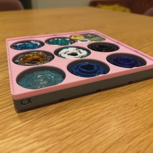 May include: A square pink and grey storage tray featuring nine circular compartments, each holding a different coloured spinning top toy. The tray has the letters CX printed in black on the front side, and the collection includes blue, gold, white, and black tops.