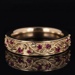 May include: A gold band ring featuring an intricate engraved leaf and vine pattern across the outer surface. The design is accented with multiple small, square-cut, deep red rubies embedded within the textured metal foliage.
