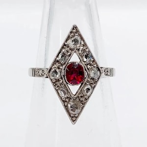 May include: A silver-toned ring featuring a central oval-shaped red gemstone set within a diamond-shaped frame. The frame is decorated with multiple small, clear, faceted stones arranged in a geometric pattern around the center stone.