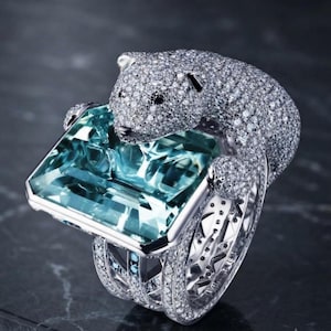 May include: A silver-toned ring featuring a large, rectangular, light blue gemstone held by a sculpted figure of a bear. The bear and the band are covered in small, clear, sparkling stones. The ring has a wide, multi-layered band design.