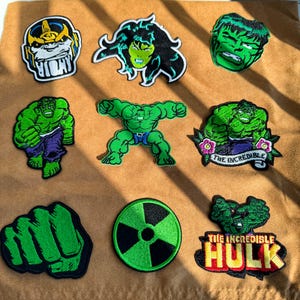 Peut inclure: Un ensemble de neuf écussons brodés représentant divers motifs de personnages de bandes dessinées verts, dont un poing, un symbole de radiation et le texte THE INCREDIBLE HULK, disposés en grille sur un tissu beige avec des ombres.