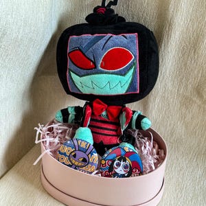 May include: A plush character with a black television-shaped head, red eyes, and a wide green grin. It wears a red bowtie and a red and black striped outfit. Two circular pins featuring cartoon characters are placed in front of the plush.