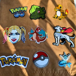 May include: A collection of nine embroidered patches featuring various characters and logos, including the Pokemon GO logo, Bulbasaur, Charmander, Harley Quinn, Charizard, a dog with a red cape, the Pokemon logo, a Poke Ball, and Vaporeon.