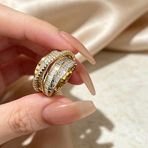 May include: A gold-toned ring featuring a multi-layered crossover design. The band is encrusted with rows of small, clear, sparkling stones and a central section of baguette-cut stones, creating a layered, textured appearance on the finger.