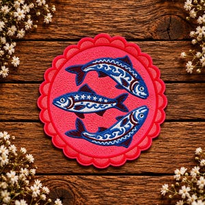 May include: A circular embroidered patch with a scalloped edge in bright pink, featuring three blue fish arranged in a circular pattern. Each fish is detailed with white star and wave motifs, dark blue fins, and a white eye, all stitched onto fabric.