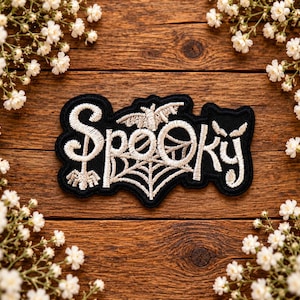 May include: A black embroidered patch featuring the word Spooky in white thread. The letter O is replaced by a spider web design, and a small bat is positioned above the web. The patch has a black border and a decorative spider at the end.