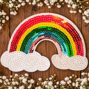 May include: A rainbow-shaped patch featuring horizontal rows of red, orange, yellow, green, blue, and purple sequins. The arch is flanked by two white clouds covered in white sequins, with a white felt border outlining the entire shape.