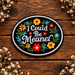 May include: An oval-shaped black embroidered patch featuring the white text I Could Be Meaner in the centre. The text is surrounded by colourful embroidered flowers in shades of red, yellow, and light blue, along with green leaves and stems.