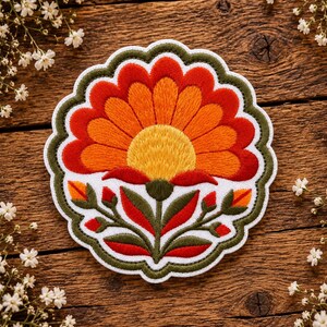 May include: A circular embroidered patch featuring a stylised flower with a bright yellow centre, orange petals, and a dark red outer layer. The design includes green leaves and stems on a white background, finished with a scalloped green border.