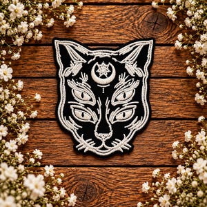 May include: A black embroidered patch featuring a cat head with four eyes and a crescent moon symbol on the forehead. The design is rendered in white thread with intricate line work, showcasing a symmetrical feline face with whiskers.