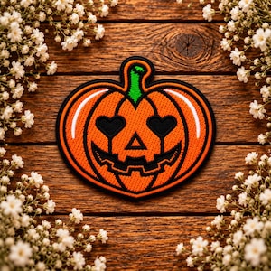 May include: An embroidered patch shaped like a pumpkin with a bright orange body, a green stem, and a black border. The pumpkin features a carved face with two heart-shaped eyes, a triangular nose, and a jagged, toothy grin in black thread.