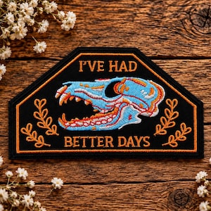 May include: A black embroidered patch with an orange border featuring a light blue animal skull graphic in the centre. The text "I'VE HAD" is stitched in orange above the skull, and "BETTER DAYS" is stitched in orange below the skull.