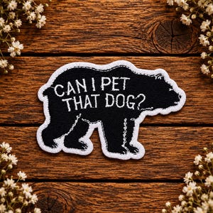 May include: A black embroidered patch in the shape of a bear with a white border. The text "CAN I PET THAT DOG?" is stitched in white thread across the centre of the bear's body, featuring a clear, bold font style against the black background.