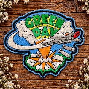 May include: An embroidered patch featuring the text GREEN DAY in green letters with yellow outlines. The design includes a grey fighter jet, two grey bombs, white clouds, and an orange explosion, all set against a bright blue background.
