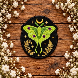 May include: A circular embroidered patch featuring a bright green luna moth with purple eye spots on a black background. Above the moth is a yellow crescent moon, and below are golden fern-like botanical sprigs and small four-pointed stars.