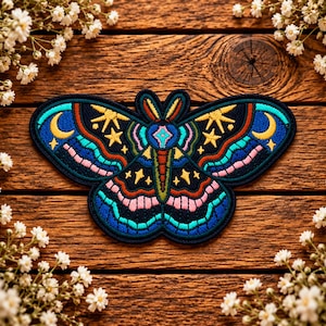 May include: An embroidered patch shaped like a moth with a dark blue background. The design features yellow crescent moons and stars, with intricate patterns in shades of bright blue, pink, and red thread throughout the wings and body.