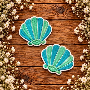 May include: Two embroidered seashell patches featuring alternating vertical stripes of teal and light green thread. Each shell has a scalloped edge and a rounded base, finished with a white stitched border around the entire perimeter of the design.