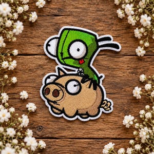 May include: An embroidered patch featuring a green cartoon character with large white eyes and a small red tongue riding on the back of a tan pig with a single large white eye and a curled tail, all outlined in a thick white border.
