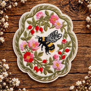 May include: An embroidered patch featuring a central bumblebee surrounded by a wreath of pink and red flowers, green leaves, and small red berries. The patch has a scalloped edge with a green border on a cream-coloured fabric background.