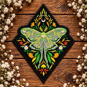 May include: A diamond-shaped black fabric patch featuring intricate embroidery of a light green moth with long tails, surrounded by yellow, orange, and white flowers, green leaves, and a small white crescent moon symbol at the top centre.