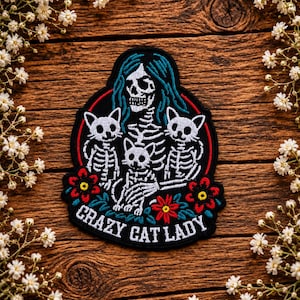 May include: A black embroidered patch featuring a skeleton figure with teal hair holding three small cat skeletons. The design includes red and yellow flowers, green leaves, and the white text CRAZY CAT LADY at the bottom, all enclosed within a red circular border.